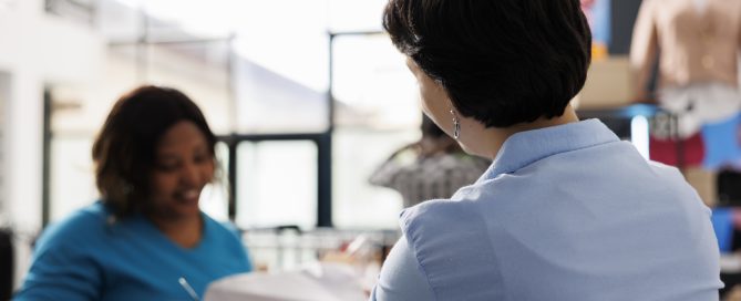Cliente afro-américaine achetant des vêtements de cérémonie, tenant plusieurs chemises dans une boutique moderne. Élégante, elle se tient au comptoir et discute des matières des articles avec un vendeur. Concept de mode