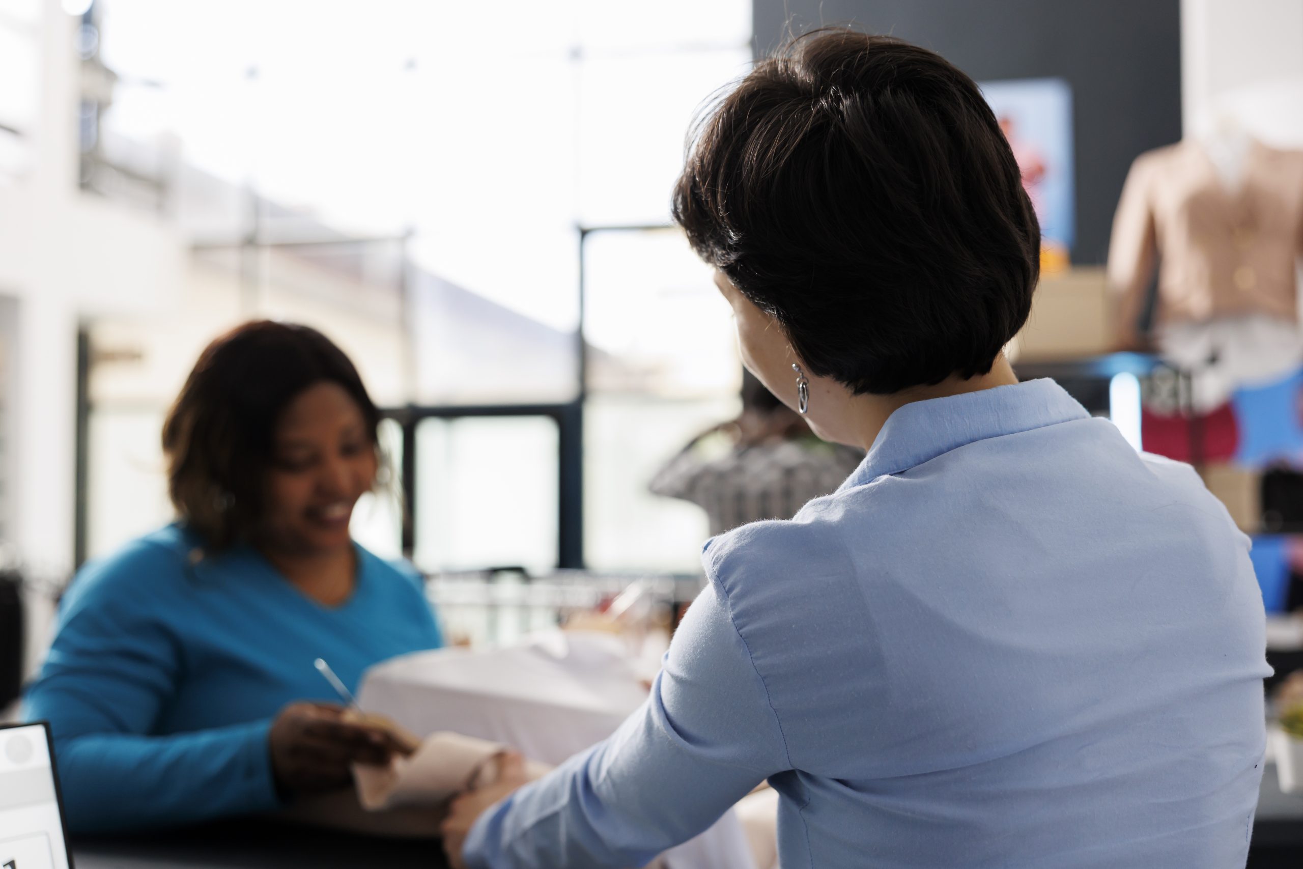 Cliente afro-américaine achetant des vêtements de cérémonie, tenant plusieurs chemises dans une boutique moderne. Élégante, elle se tient au comptoir et discute des matières des articles avec un vendeur. Concept de mode